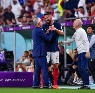 Soccer players shake hands on the sideline during a FIFA World Cup match (Qatar 2022), with a cameraman filming nearby.