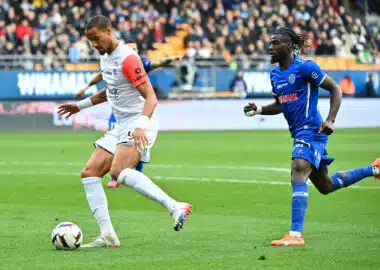 Two soccer players, one in white with a red sleeve and one in blue, race for the ball on a green pitch with a crowded stadium backdrop.