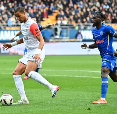 Two soccer players, one in white with a red sleeve and one in blue, race for the ball on a green pitch with a crowded stadium backdrop.