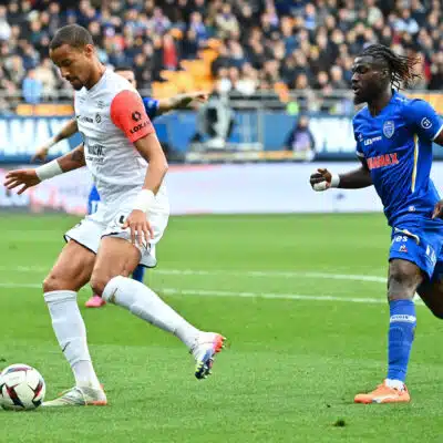 Two soccer players, one in white with a red sleeve and one in blue, race for the ball on a green pitch with a crowded stadium backdrop.
