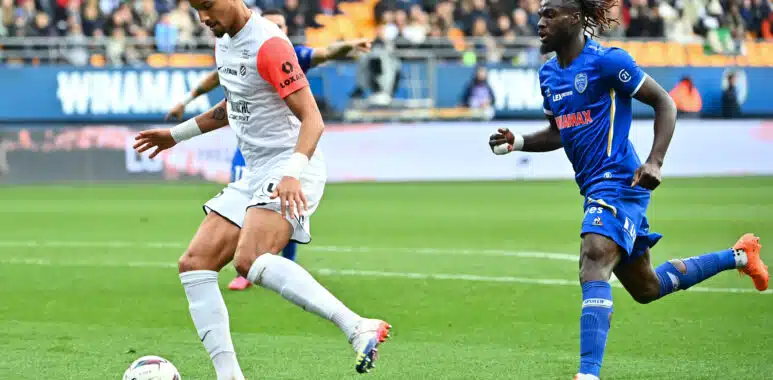 Christopher Jullien ESTAC Troyes v Montpellier Herault SC - Ligue 1 Uber Eats | AllezPaillade.com Two soccer players, one in white with a red sleeve and one in blue, race for the ball on a green pitch with a crowded stadium backdrop.