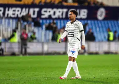 Soccer player in a white patterned jersey walking on a grass pitch with blurred stadium seating in the background.