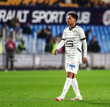 Soccer player in a white patterned jersey walking on a grass pitch with blurred stadium seating in the background.