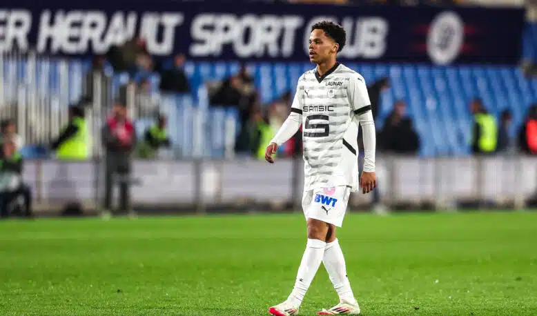 Soccer player in a white patterned jersey walking on a grass pitch with blurred stadium seating in the background.