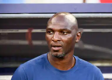 Close-up of a man wearing a blue shirt, looking to the right, with blurred stadium seating behind him.