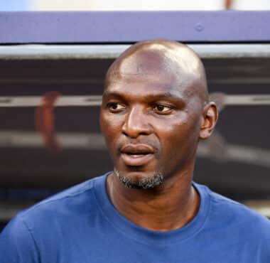 Close-up of a man wearing a blue shirt, looking to the right, with blurred stadium seating behind him.