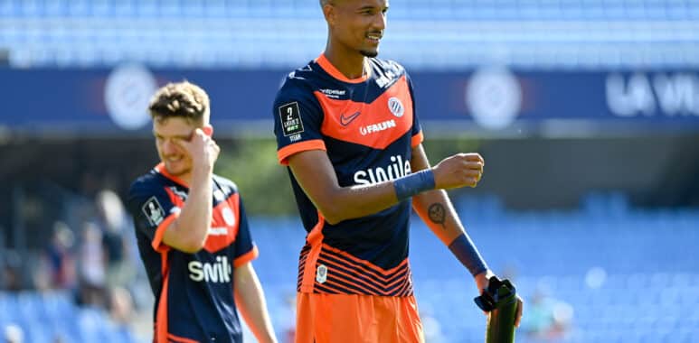 Soccer player in navy and orange kit celebrates on the field, with a teammate in the background under stadium lights.
