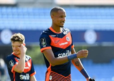 Soccer player in navy and orange kit celebrates on the field, with a teammate in the background under stadium lights.