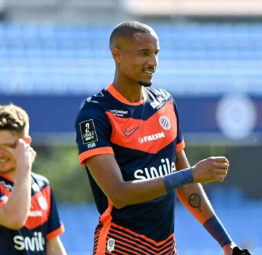 Soccer player in navy and orange kit celebrates on the field, with a teammate in the background under stadium lights.