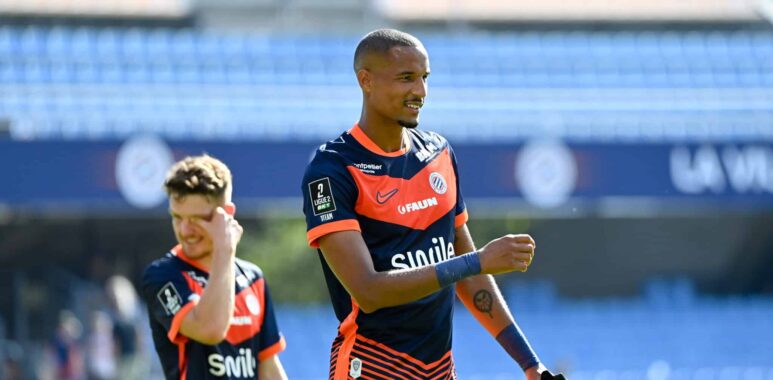 Soccer player in navy and orange kit celebrates on the field, with a teammate in the background under stadium lights.