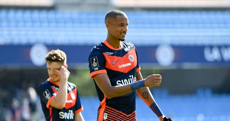 Soccer player in navy and orange kit celebrates on the field, with a teammate in the background under stadium lights.