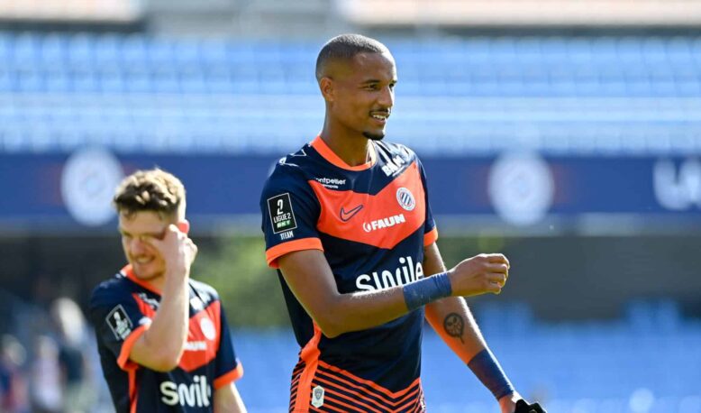 Soccer player in navy and orange kit celebrates on the field, with a teammate in the background under stadium lights.