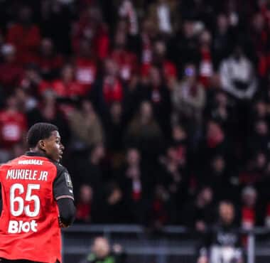 Soccer player Mukiele Jr. in a red jersey with number 65, seen from behind, on a crowded stadium.