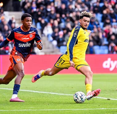 Soccer players in yellow and blue uniforms chase the ball on a green field with a crowd in the stands watching a match ongoing behind them.