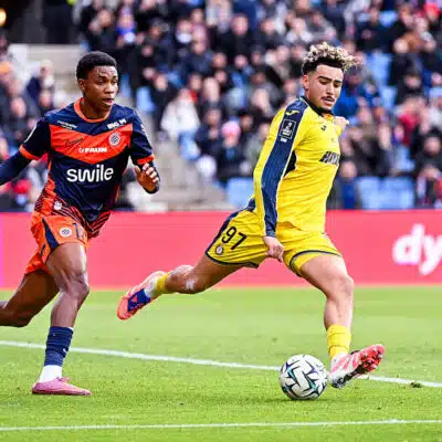 Soccer players in yellow and blue uniforms chase the ball on a green field with a crowd in the stands watching a match ongoing behind them.