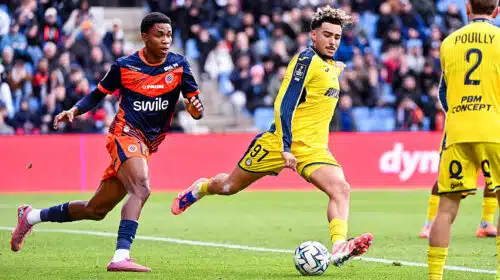 Soccer players in yellow and blue uniforms chase the ball on a green field with a crowd in the stands watching a match ongoing behind them.