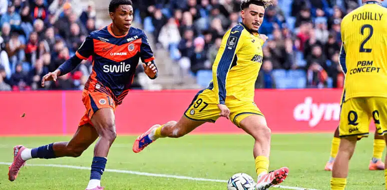 Montpellier Herault SC v Pau FC - Ligue 2 BKT 20252026 | AllezPaillade.com Soccer players in yellow and blue uniforms chase the ball on a green field with a crowd in the stands watching a match ongoing behind them.