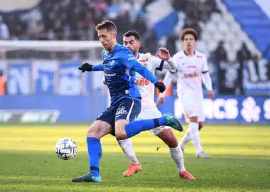 Benet | AllezPaillade.com Soccer players compete for the ball on a sunny field; a blue-team player in front, white-team defender close behind.