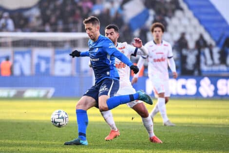 Soccer players compete for the ball on a sunny field; a blue-team player in front, white-team defender close behind.