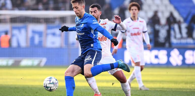 Benet | AllezPaillade.com Soccer players compete for the ball on a sunny field; a blue-team player in front, white-team defender close behind.