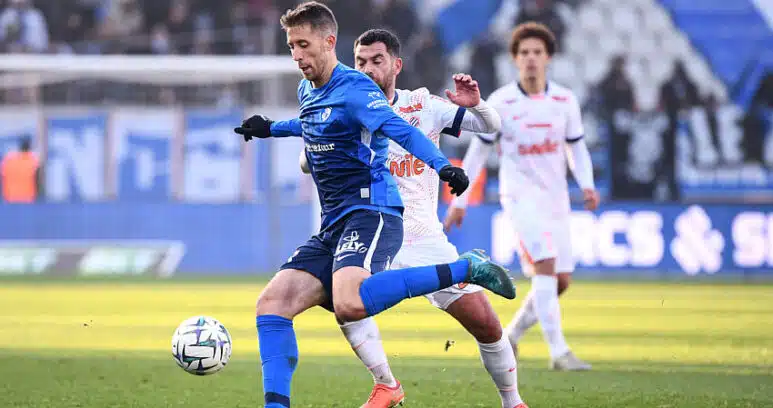 Soccer players compete for the ball on a sunny field; a blue-team player in front, white-team defender close behind.