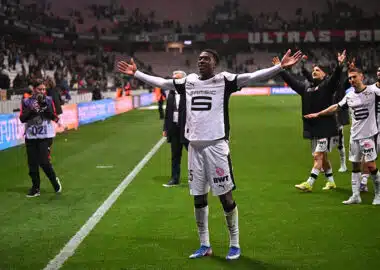 Mukiele | AllezPaillade.com Soccer player in white uniform raises arms in celebration near the sideline as teammates applaud in the background on a green pitch