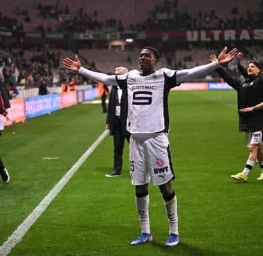 Soccer player in white uniform raises arms in celebration near the sideline as teammates applaud in the background on a green pitch