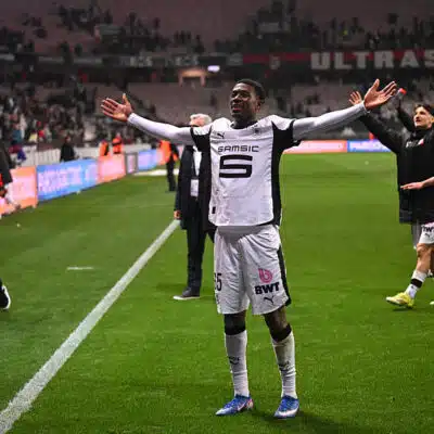 Soccer player in white uniform raises arms in celebration near the sideline as teammates applaud in the background on a green pitch