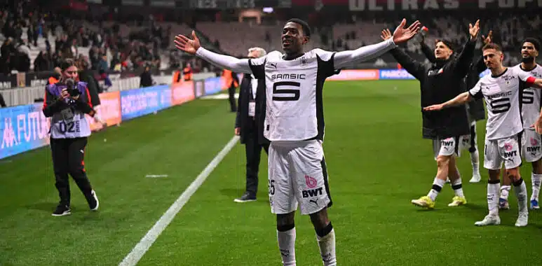 Soccer player in white uniform raises arms in celebration near the sideline as teammates applaud in the background on a green pitch