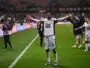 Soccer player in white uniform raises arms in celebration near the sideline as teammates applaud in the background on a green pitch