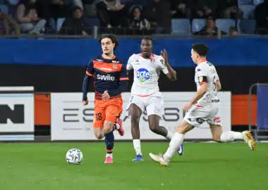Soccer players in orange and white jerseys chase a ball on a green field during a match, with spectators in the stands behind them.