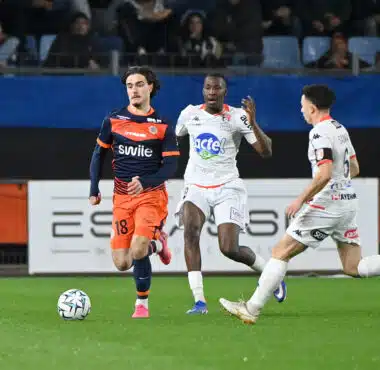 Soccer players in orange and white jerseys chase a ball on a green field during a match, with spectators in the stands behind them.