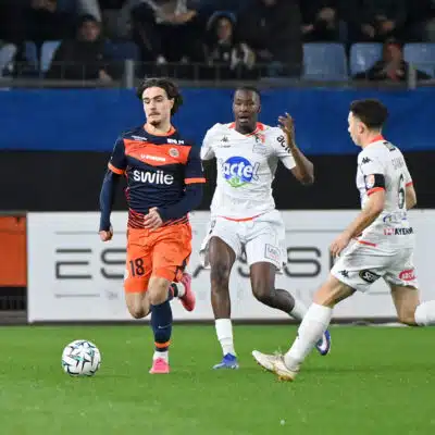 Soccer players in orange and white jerseys chase a ball on a green field during a match, with spectators in the stands behind them.