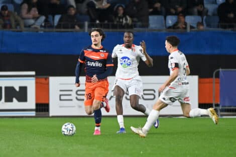 Soccer players in orange and white jerseys chase a ball on a green field during a match, with spectators in the stands behind them.