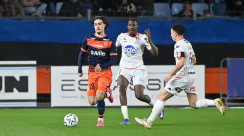 Soccer players in orange and white jerseys chase a ball on a green field during a match, with spectators in the stands behind them.