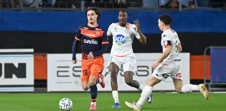 Soccer players in orange and white jerseys chase a ball on a green field during a match, with spectators in the stands behind them.