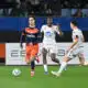 Soccer players in orange and white jerseys chase a ball on a green field during a match, with spectators in the stands behind them.