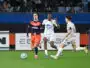 Soccer players in orange and white jerseys chase a ball on a green field during a match, with spectators in the stands behind them.