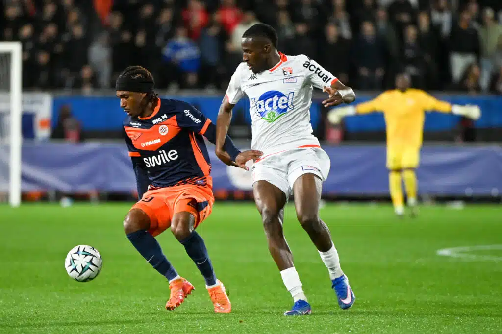 Two footballers chase the ball during a match: navy-and-orange kit versus white kit, with a yellow-jersey keeper visible in the background.