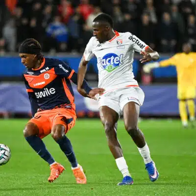 Two footballers chase the ball during a match: navy-and-orange kit versus white kit, with a yellow-jersey keeper visible in the background.