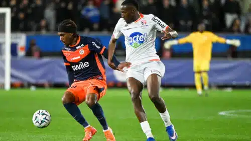 Two footballers chase the ball during a match: navy-and-orange kit versus white kit, with a yellow-jersey keeper visible in the background.