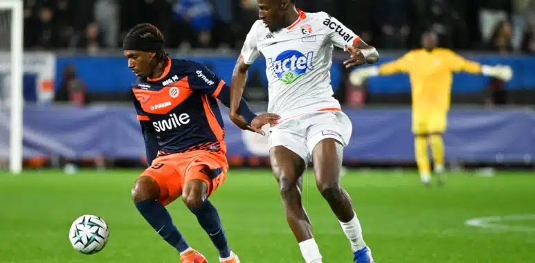 Two footballers chase the ball during a match: navy-and-orange kit versus white kit, with a yellow-jersey keeper visible in the background.