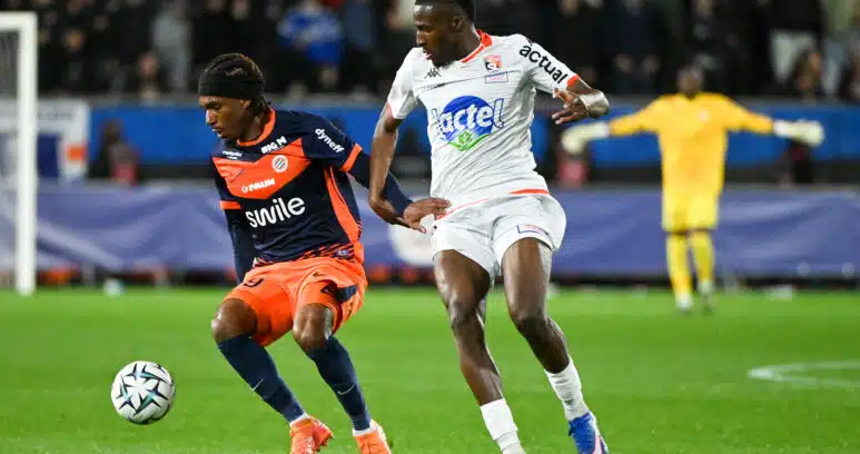 Two footballers chase the ball during a match: navy-and-orange kit versus white kit, with a yellow-jersey keeper visible in the background.