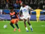 Two footballers chase the ball during a match: navy-and-orange kit versus white kit, with a yellow-jersey keeper visible in the background.
