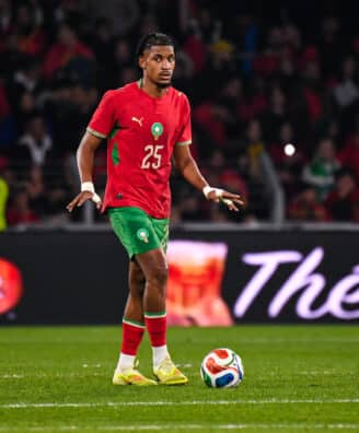 Soccer player in red jersey with green shorts dribbling the ball on a green field during a match with a crowded stadium behind him