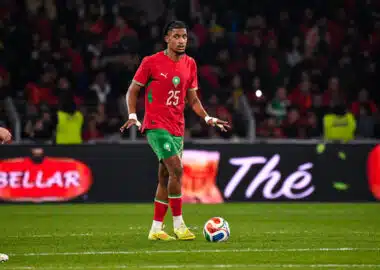 Soccer player in red jersey with green shorts dribbling the ball on a green field during a match with a crowded stadium behind him
