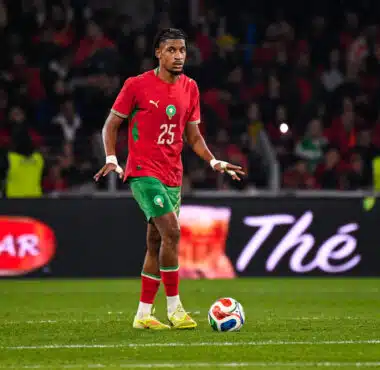 Soccer player in red jersey with green shorts dribbling the ball on a green field during a match with a crowded stadium behind him