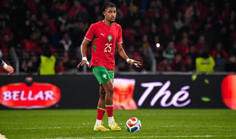 Soccer player in red jersey with green shorts dribbling the ball on a green field during a match with a crowded stadium behind him