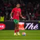 Soccer player in red jersey with green shorts dribbling the ball on a green field during a match with a crowded stadium behind him