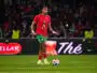 Soccer player in red jersey with green shorts dribbling the ball on a green field during a match with a crowded stadium behind him